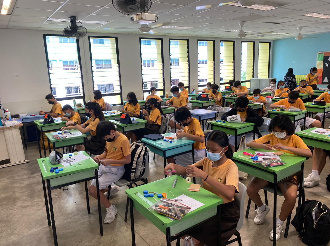 Students in yellow shirts and face masks sit at green desks working on craft projects in a bright classroom.